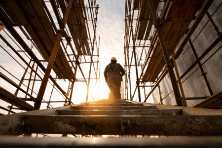 Man with hard hat on, walking up ladder in between scaffolding buildings